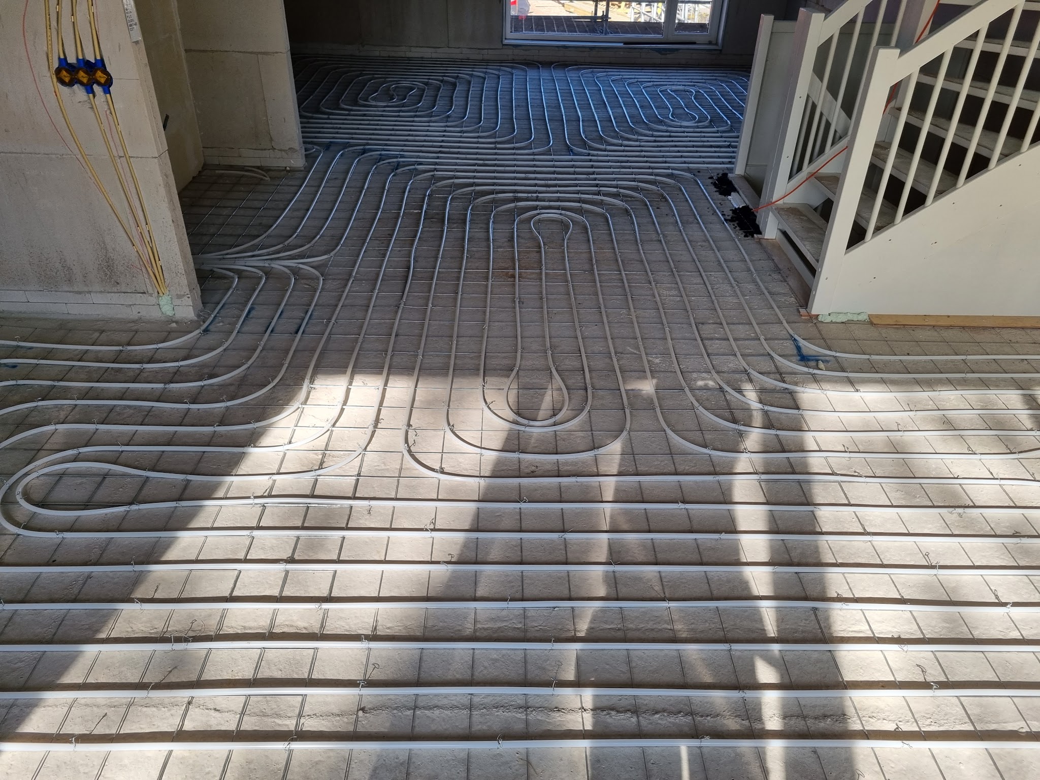 Een woonkamer met pas ingefreesde vloerverwarming in lussen, met zonlicht en een witte trap