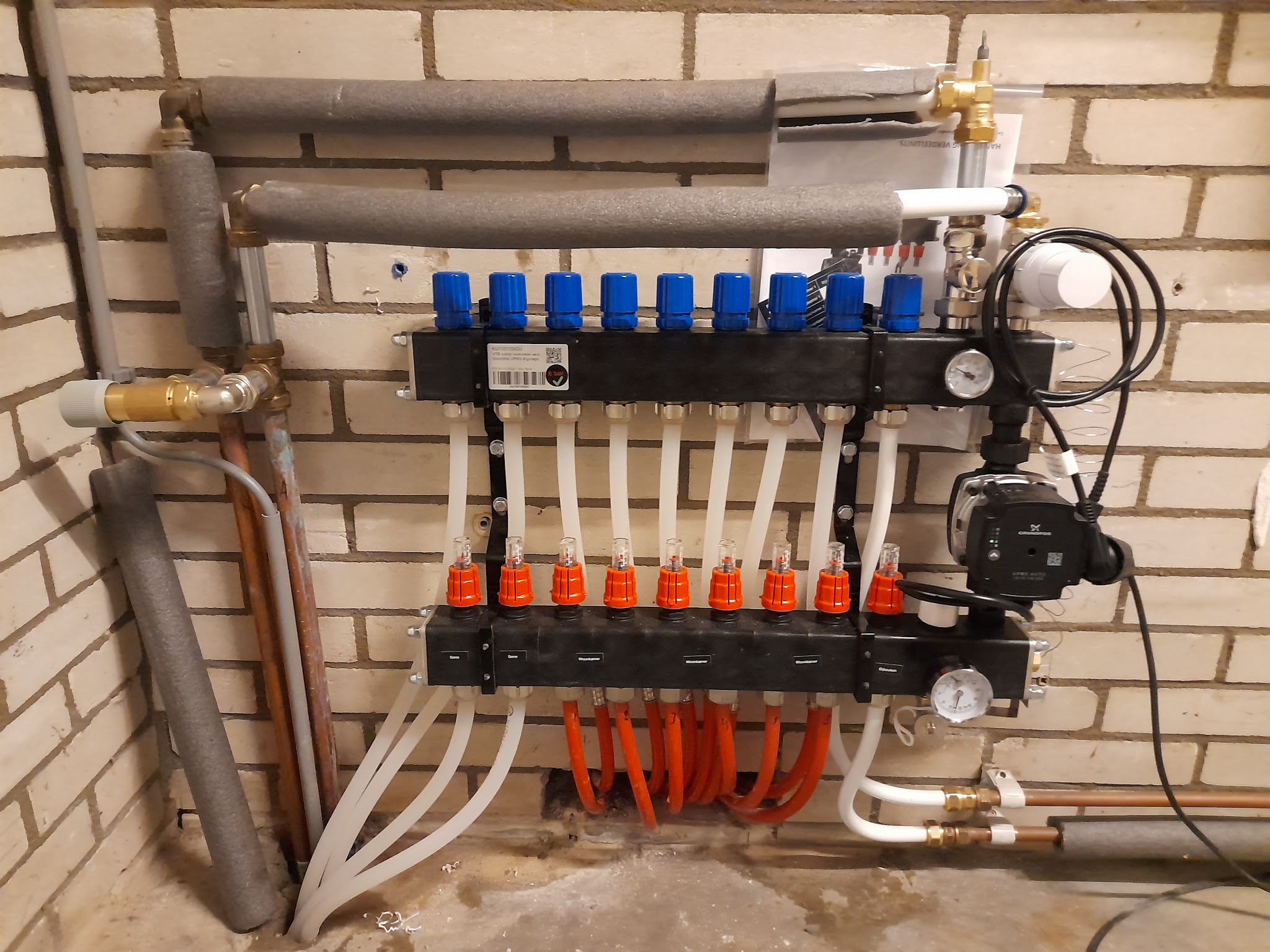 Een aangesloten vloerverwarmingsverdeler met blauwe en rode knoppen en een pomp tegen een bakstenen muur
