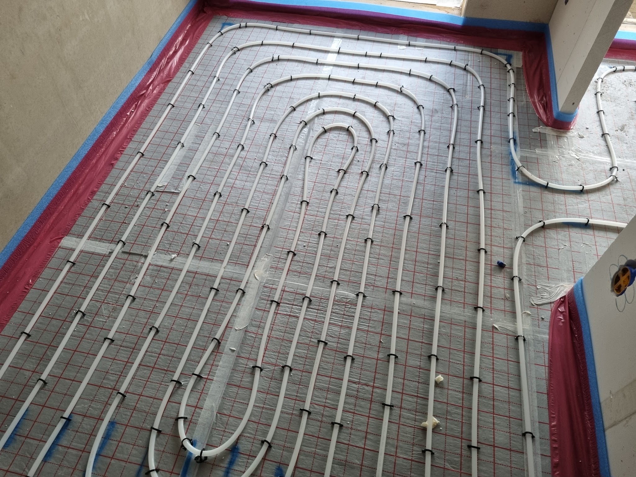 Witte vloerverwarmingsbuizen in lussen gebonden op een wapeningsnet, met rode randstrook langs de wand