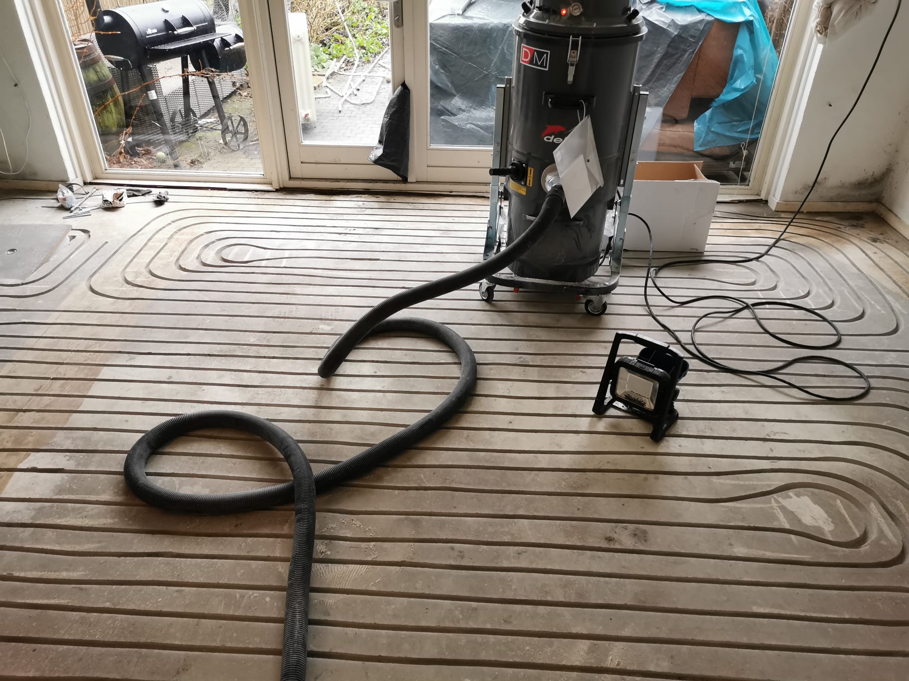Gefreesde sleuven met vloerverwarmingsbuizen in een bestaande vloer, met een bouwstofzuiger bij de tuindeuren